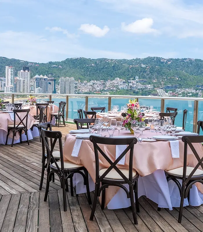 Terraza con mesas elegantes, vista al mar y edificios al fondo.