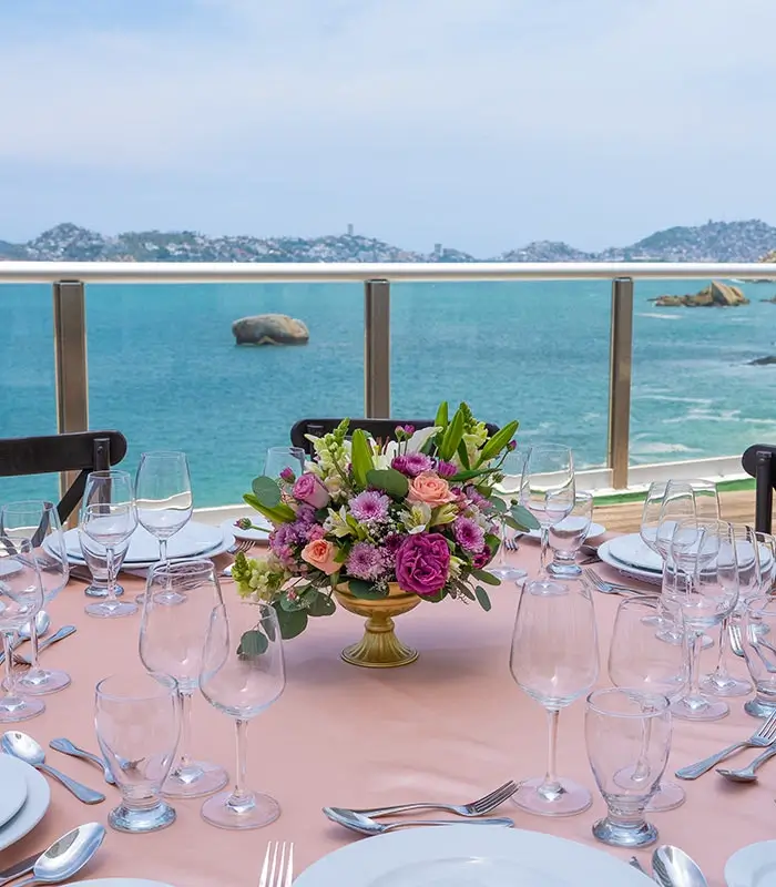 Mesa elegante con vista al mar, decorada con un centro floral y copas de vino alrededor.