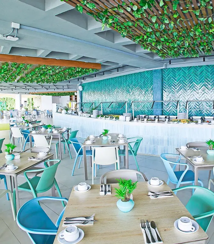 Restaurante moderno con decoraci&oacute;n verde y azul, mesas dispuestas y techo decorado con plantas.