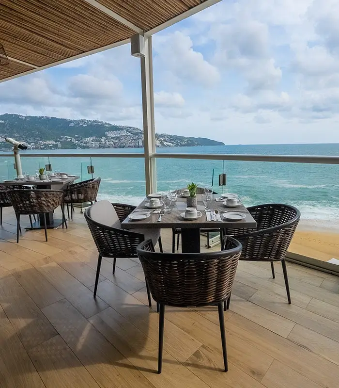 Terraza de restaurante con mesas y sillas, vista al mar y monta&ntilde;as en el fondo.