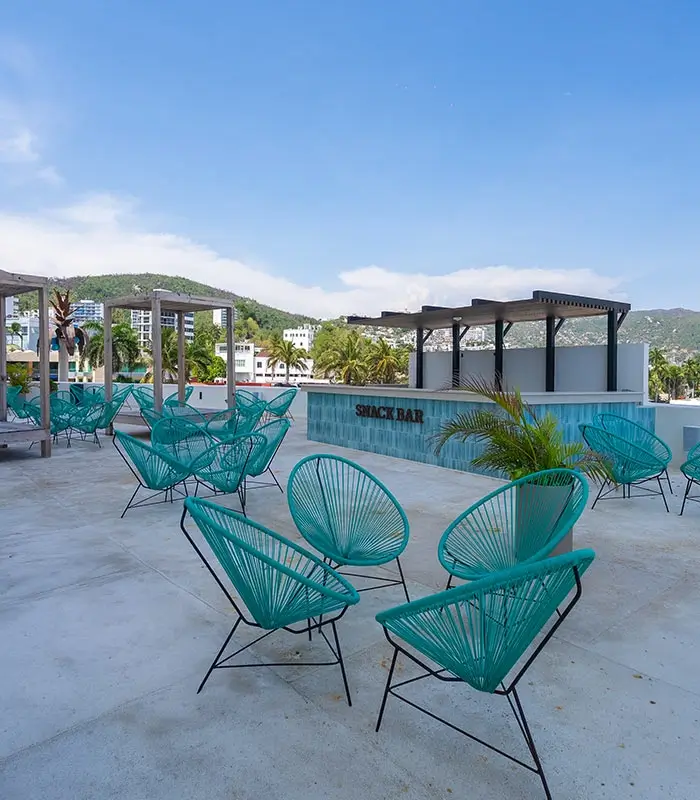Terraza al aire libre con sillas azules y un snack bar, vegetaci&oacute;n y monta&ntilde;as al fondo.