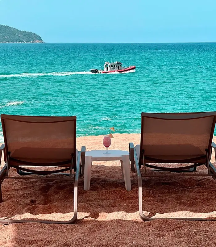 Sillas en la playa frente al mar con una bebida sobre una mesa; un bote pasa en el fondo.