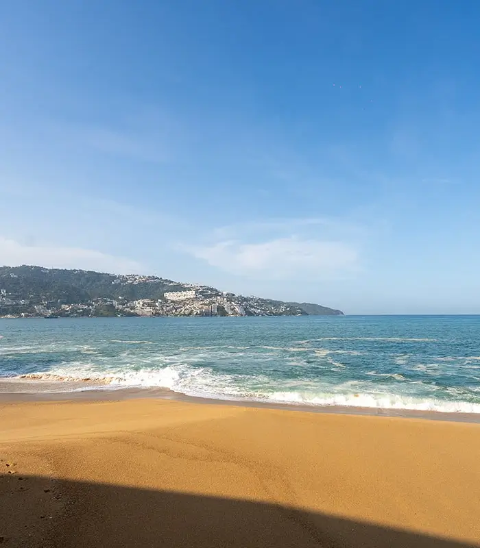 Playa con arena dorada, olas del mar y una colina con casas bajo un cielo azul claro.