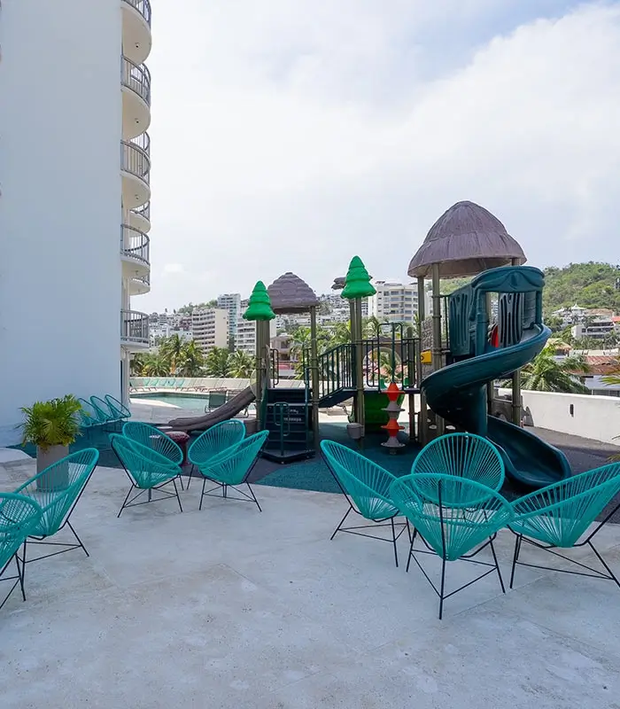 &Aacute;rea de juegos con resbaladilla y sillas turquesas en una terraza, rodeada de edificios y &aacute;rboles.
