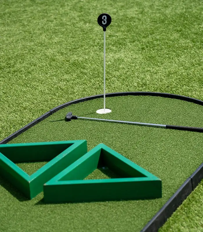 Minigolf: hoyo 3 con bander&iacute;n, obst&aacute;culos triangulares verdes y un palo de golf sobre el c&eacute;sped.