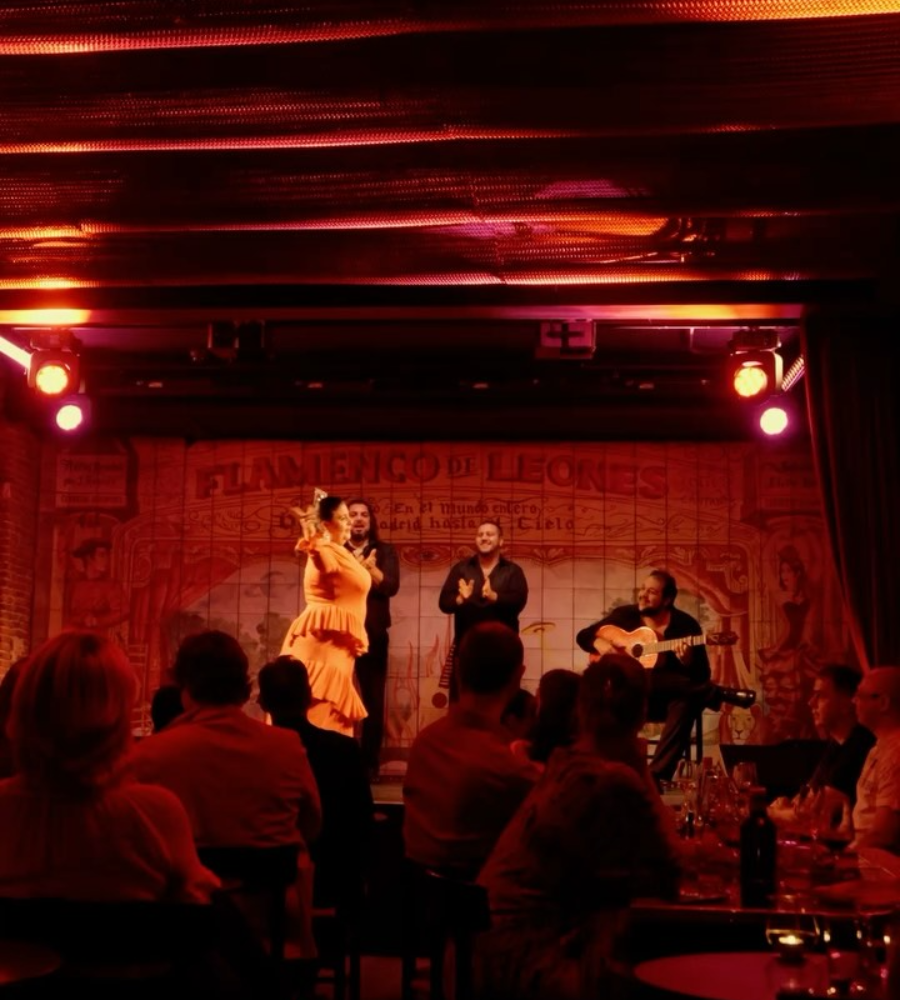Espect&aacute;culo de flamenco en tablao, con bailaora vestida de naranja, guitarrista y espectadores bajo luces c&aacute;lidas.