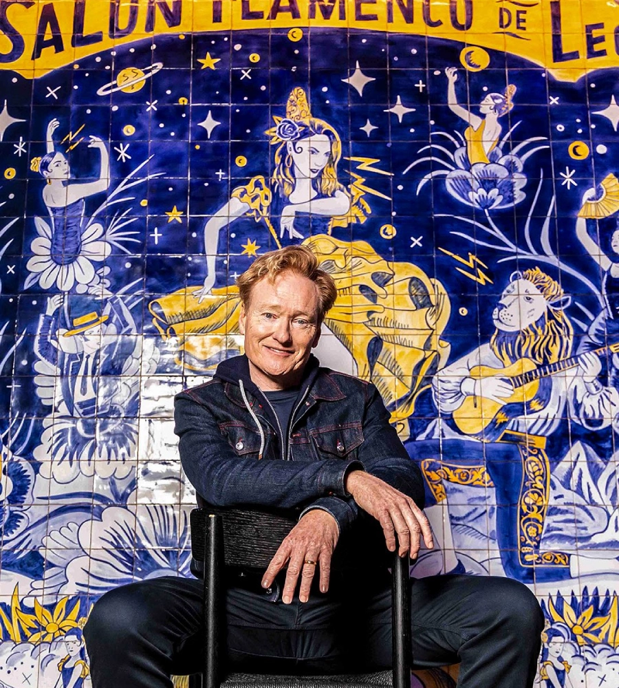 Hombre sonriente sentado frente a un mural colorido de flamenco con decoraciones de flores y animales.