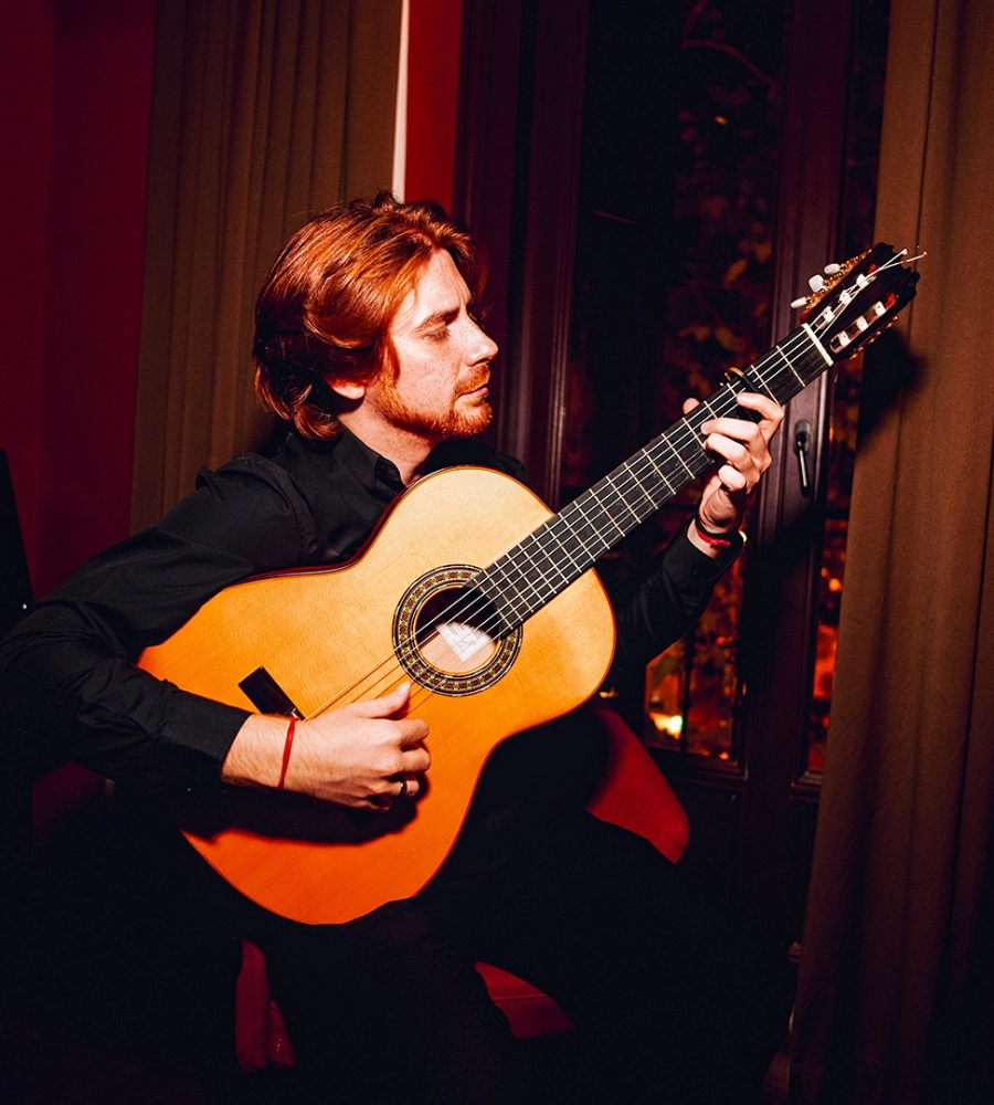Persona tocando una guitarra cl&aacute;sica, sentado junto a una ventana, en un ambiente c&aacute;lido e iluminado.