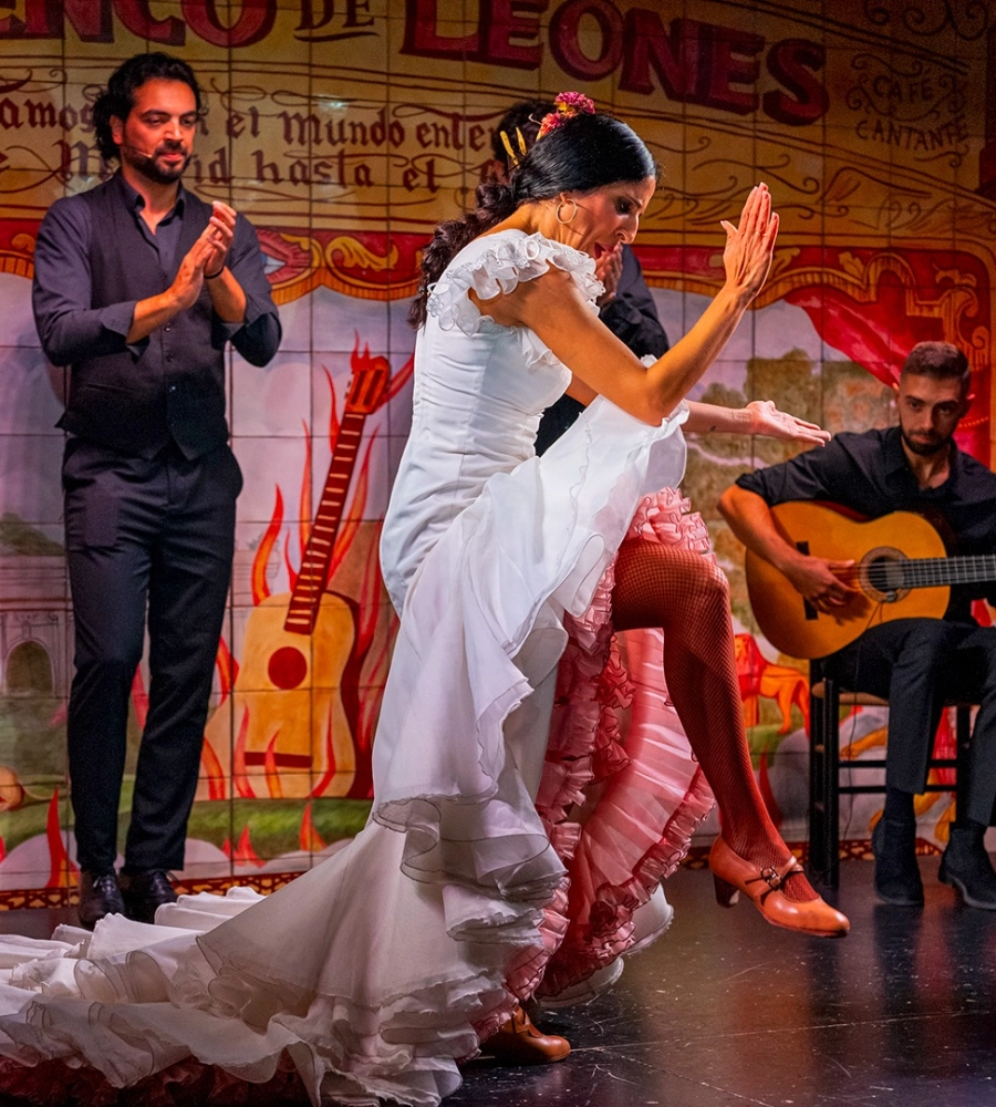 Bailarina de flamenco con vestido blanco, acompa&ntilde;ada por guitarrista y otra persona aplaudiendo sobre un escenario colorido.