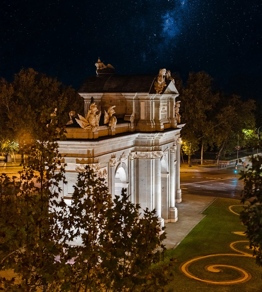 Arco de piedra iluminado de noche, rodeado de &aacute;rboles, bajo un cielo estrellado.