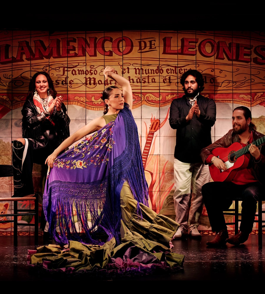 Una bailaora de flamenco con mant&oacute;n p&uacute;rpura, rodeada de m&uacute;sicos y aplausos, en un tablao decorado.
