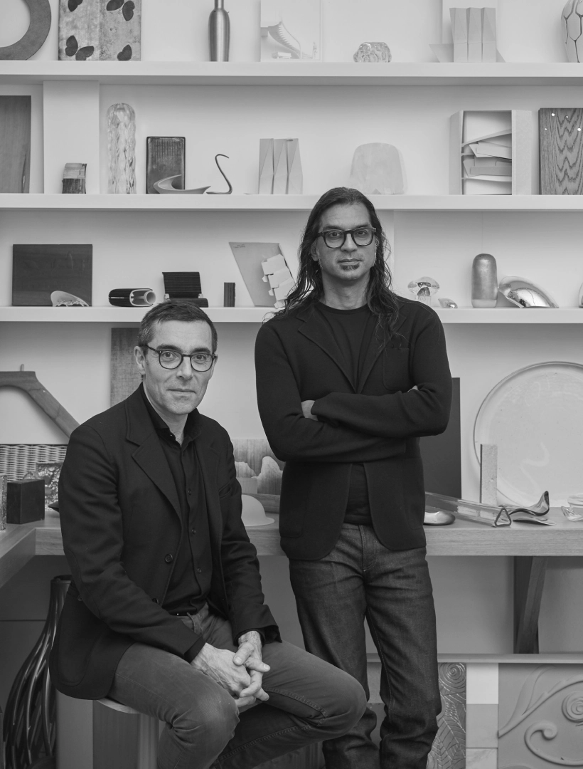 Dos hombres posan en un estudio con estanter&iacute;as llenas de objetos y esculturas abstractas en blanco y negro.