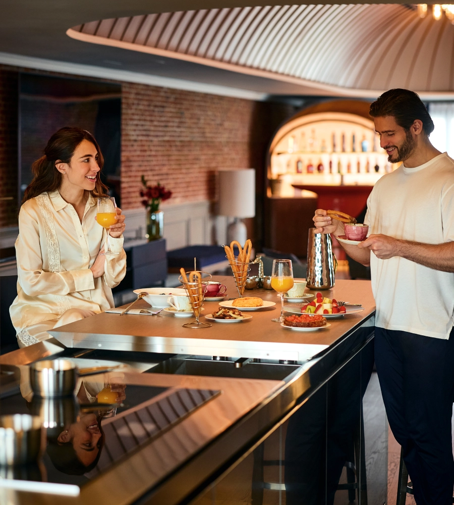 Pareja desayunando sonriendo en la cocina moderna con copas de jugo, caf&eacute; y platos con frutas, pasteles y pan.