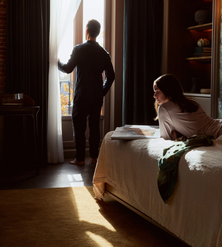 Hombre de pie mirando por la ventana y mujer acostada en la cama, ambos en una habitaci&oacute;n iluminada por la luz del sol.