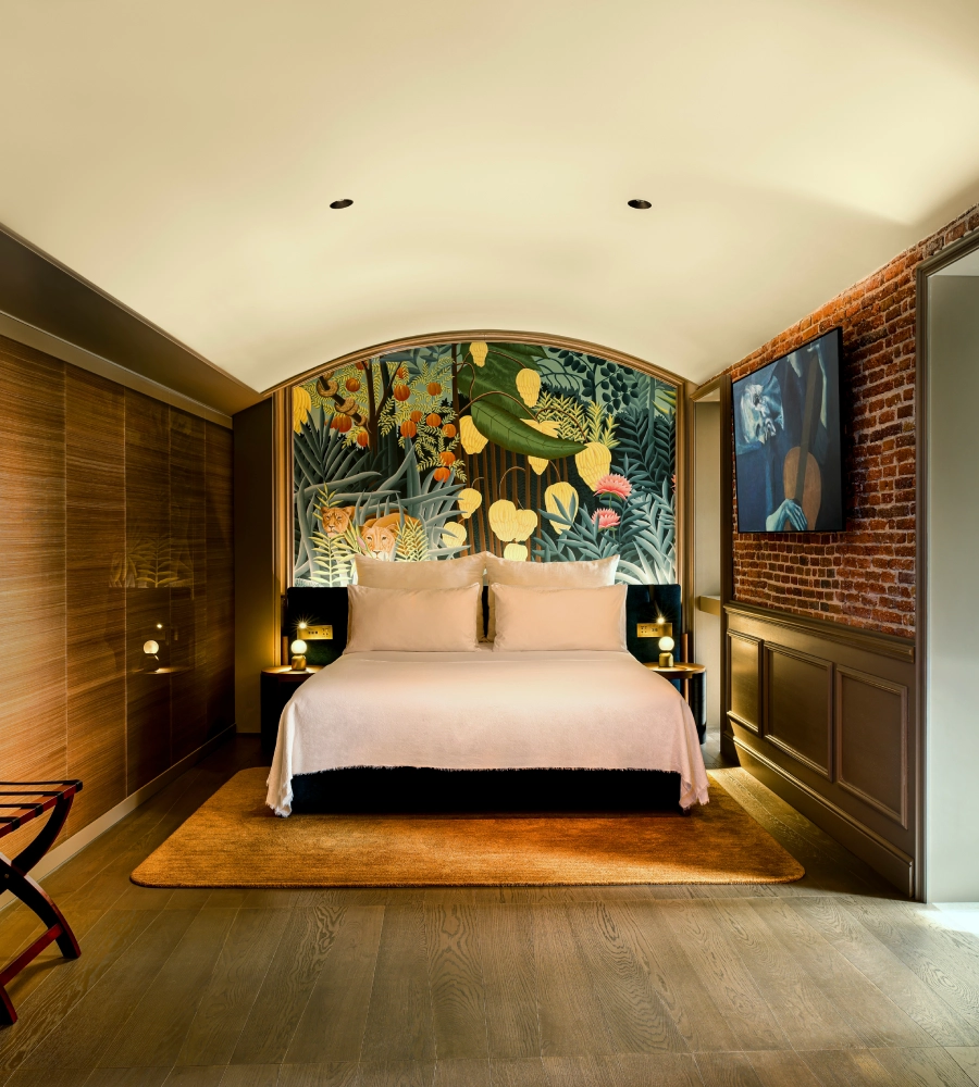 Habitaci&oacute;n moderna con cama doble blanca, mural de jungla detr&aacute;s, paredes de ladrillo y madera, y alfombra marr&oacute;n.