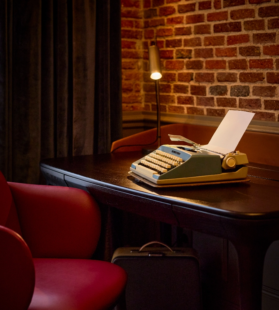 Una m&aacute;quina de escribir azul en un escritorio oscuro, junto a una l&aacute;mpara encendida y una silla roja. Fondo de ladrillos.