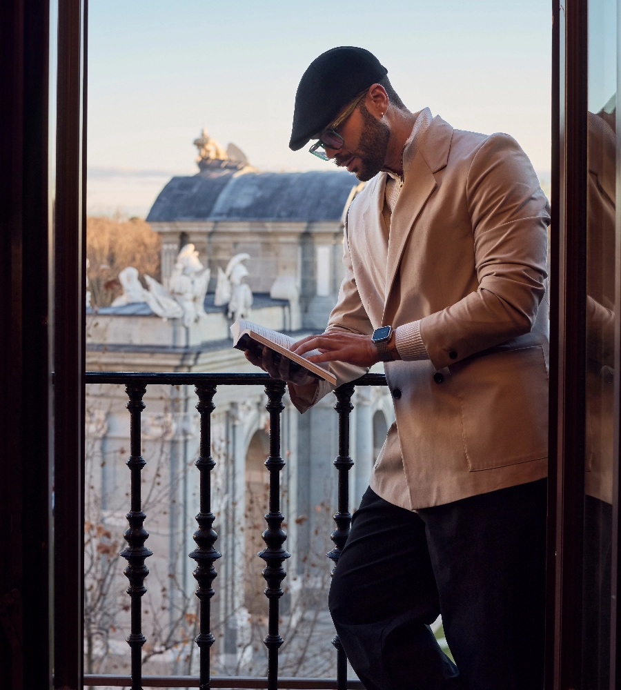 Hombre con gorra y abrigo leyendo un libro en un balc&oacute;n con esculturas y edificio hist&oacute;rico de fondo.