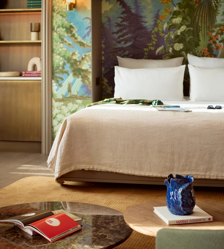 Habitaci&oacute;n con cama doble, mural de paisaje en la pared, mesa con vaso azul sobre un libro y decoraci&oacute;n moderna.