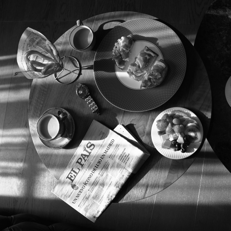 Mesa con caf&eacute;, peri&oacute;dico, reloj, bollos, frutas y tostadas, iluminada por luz natural en blanco y negro.