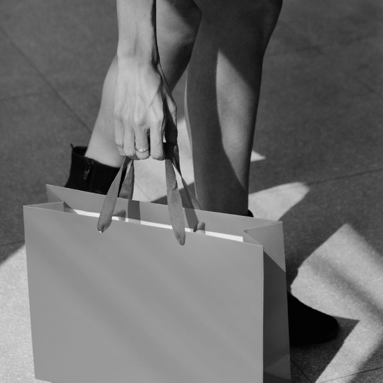 Persona con botas negras sosteniendo una bolsa de compras grande en blanco y negro, sobre un suelo con sombras.