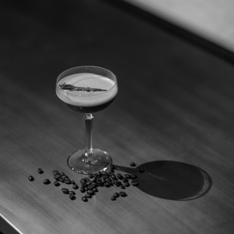 Copa de c&oacute;ctel con una rama decorativa, rodeada de granos de caf&eacute; sobre una superficie de madera. Imagen en blanco y negro.