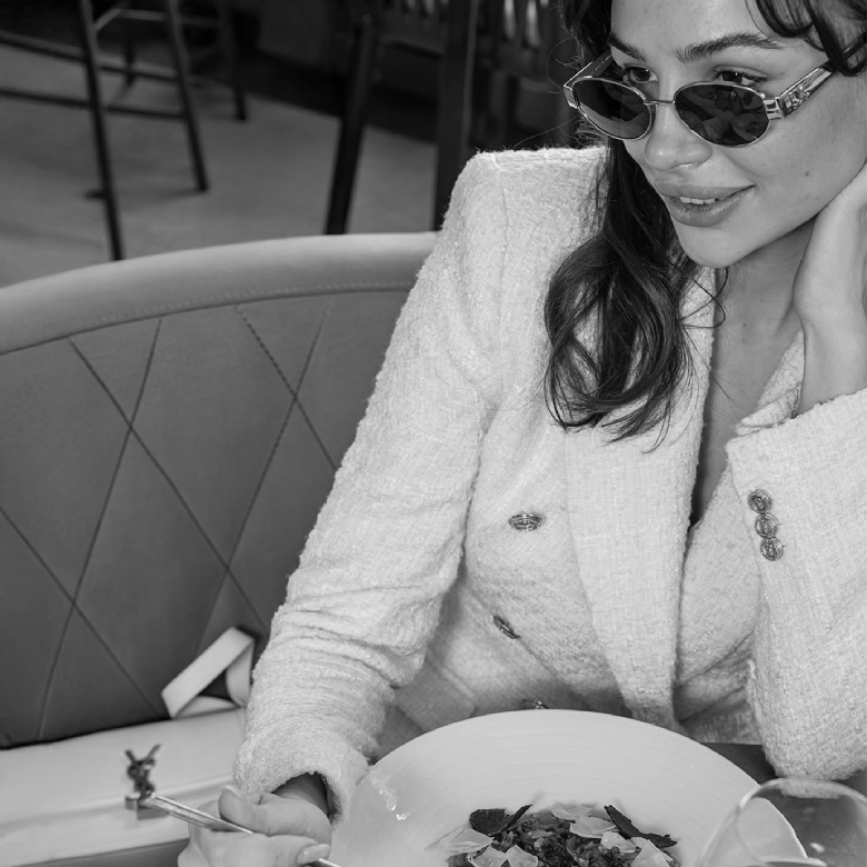 Mujer con gafas de sol y traje claro, sentada en un restaurante frente a un plato de comida. Imagen en blanco y negro.