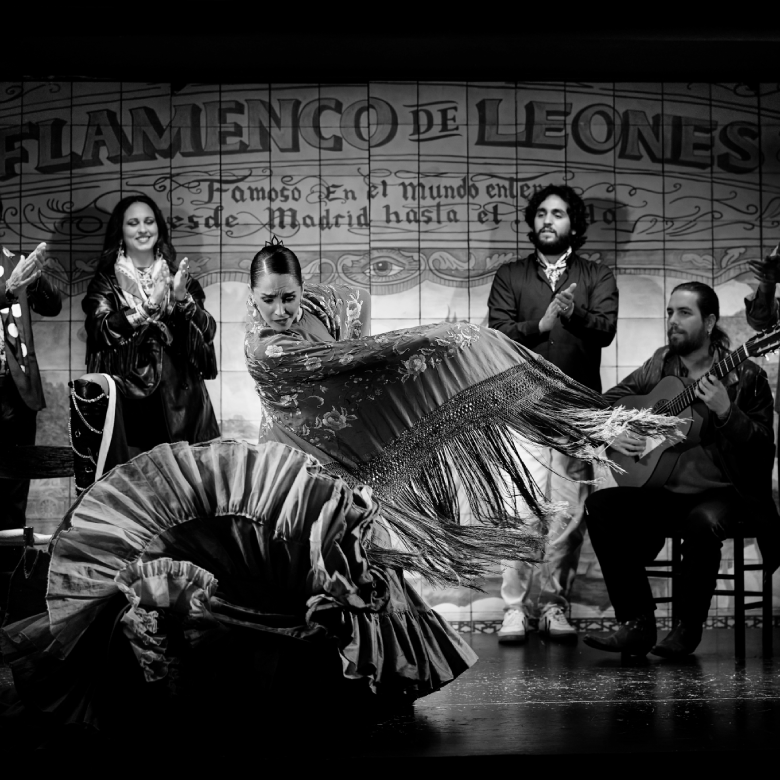 Bailarina de flamenco moviendo su vestido en un escenario, acompa&ntilde;ada de m&uacute;sicos y personas aplaudiendo. Blanco y negro.