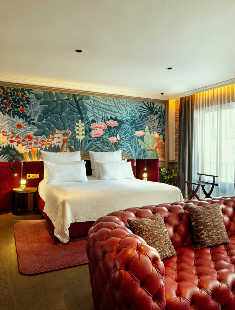 Habitaci&oacute;n moderna con cama doble, mural de jungla, sof&aacute; rojo de cuero y luz natural filtrando por cortinas claras.