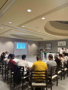 Persona dando presentaci&oacute;n en sala, p&uacute;blico sentado frente a pantalla con diapositiva.