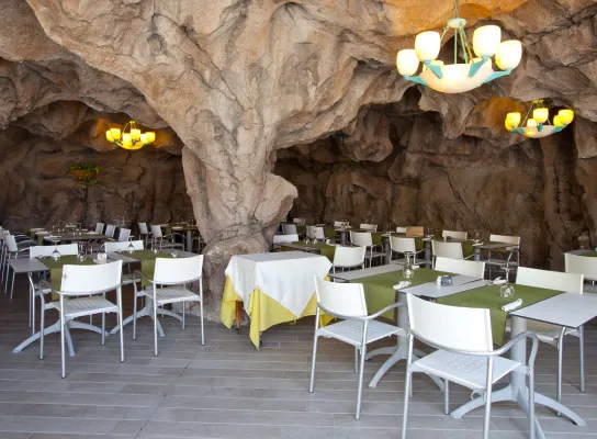 Restaurante en cueva con mesas y sillas blancas, adornado con lámparas y manteles verdes.