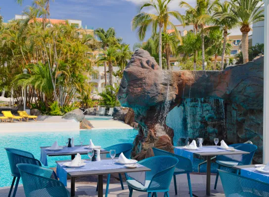 Restaurante al aire libre junto a una piscina, rodeado de palmeras, con mesas y sillas azules.