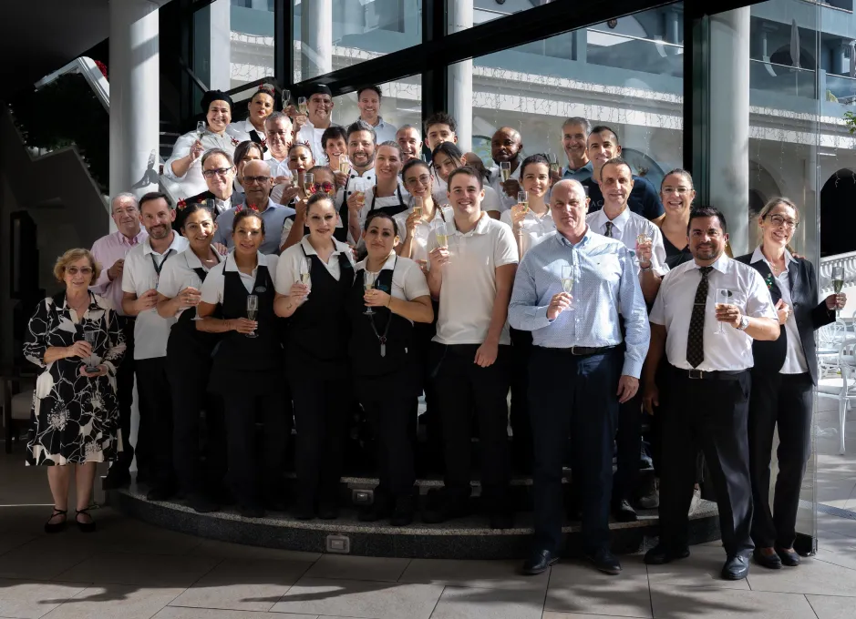 Grupo de personas celebrando con copas, posan en escaleras junto a una ventana grande.