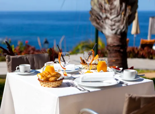 Mesa con desayuno servido junto al mar, incluye jugo, café y croissants.