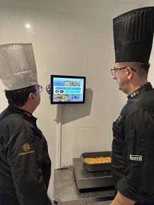 Dos chefs observan una pantalla en una cocina, junto a una bandeja de comida sobre la mesa.