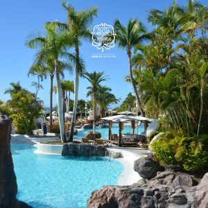 Piscina rodeada de palmeras y rocas en un hotel tropical con cielo despejado.