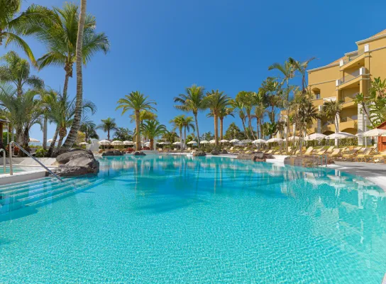 Piscina rodeada de palmeras junto a un hotel bajo un cielo despejado.
