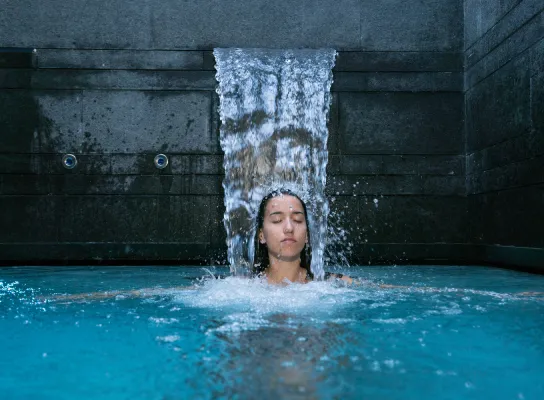 Persona bajo una cascada en una piscina cerrando los ojos, rodeada de paredes de piedra oscura.