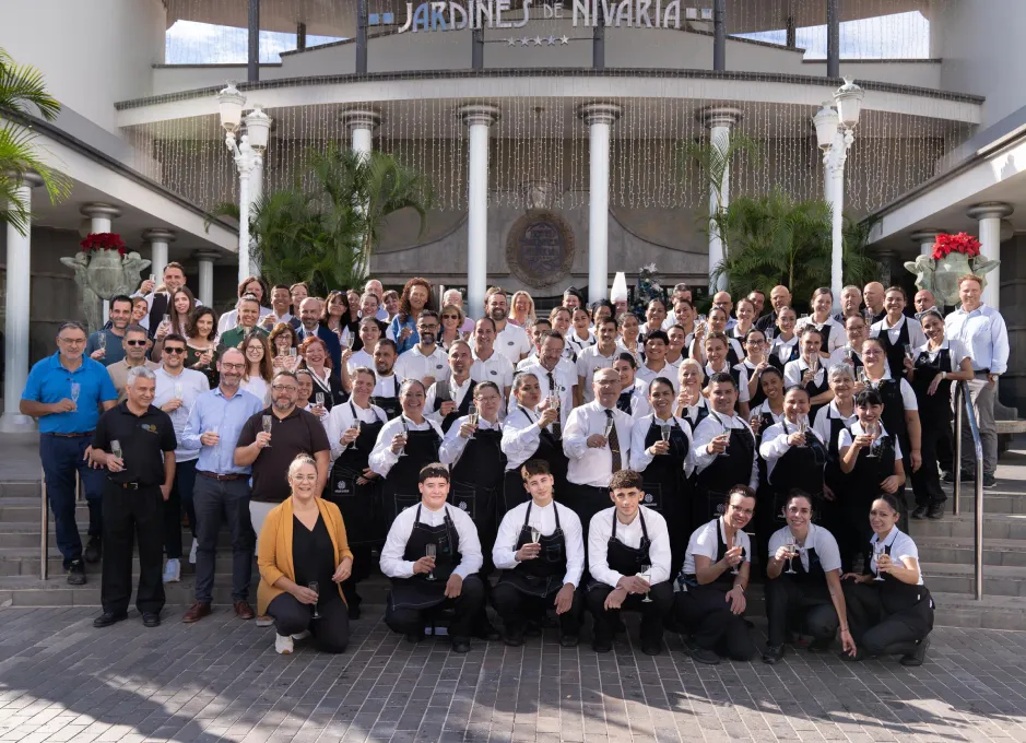 Un grupo numeroso de personas sonríe en la entrada de un edificio con la fachada "Jardines de Nivaria".