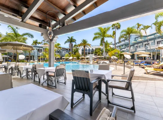 Terraza de restaurante junto a una piscina con palmeras y tumbonas en un ambiente soleado.