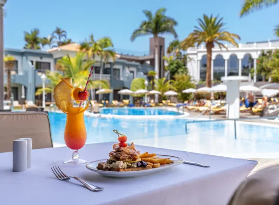 Plato de comida y cóctel sobre mesa, junto a piscina en un complejo turístico con palmeras.