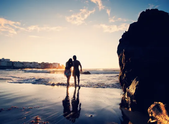 Pareja de espaldas caminando en la playa al atardecer, con reflejos en el agua.