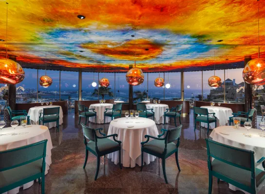 Restaurante con techo colorido, mesas redondas, sillas turquesa y vista panorámica al exterior.