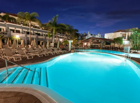Piscina iluminada por la noche con tumbonas y palmeras alrededor en un complejo turístico.
