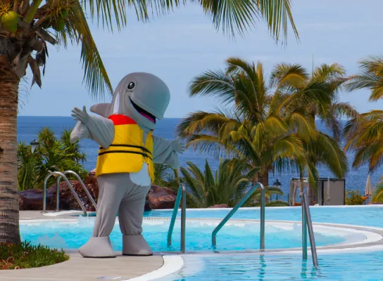 Personaje disfrazado de cetáceo con chaleco frente a una piscina, palmeras y mar de fondo.