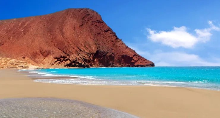 Playa con acantilado rojo, arena dorada y mar azul bajo un cielo despejado.
