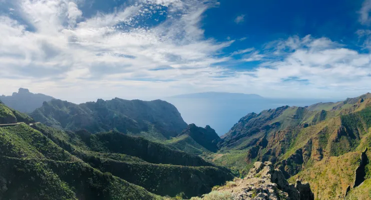 Montañas verdes bajo un cielo azul con nubes, junto a un horizonte oceánico lejano.
