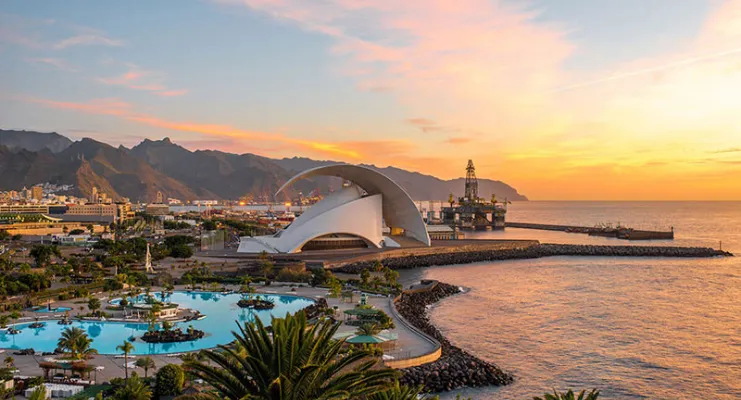 Auditorio moderno frente al mar al atardecer, con montañas y piscina en el primer plano.
