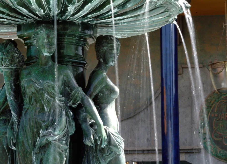 Fuente con figuras femeninas de bronce y agua cayendo sobre ellas en un ambiente exterior.