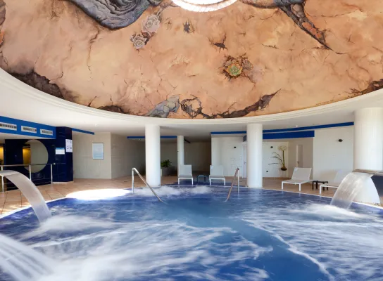 Piscina interior con cascadas y techo decorado con un mural artístico. Ambiente relajante.