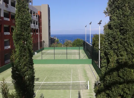 Cancha de tenis rodeada de árboles y edificios, con vista al mar al fondo.